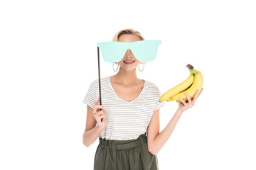 smiling young woman holding bananas and eyeglasses on party stick isolated on white