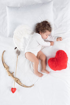 little baby with wings lying on bed with heart pillow, bow and arrow