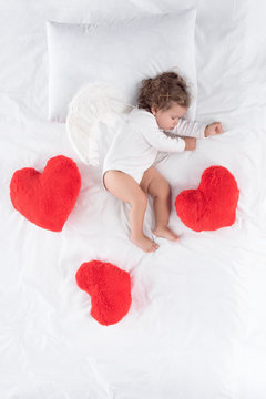 little cupid with wings lying on bed with red hearts