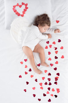 little toddler with wings sleeping on bed with red hearts