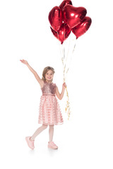 beautiful child in pink dress holding heart shaped balloons and smiling at camera isolated on white