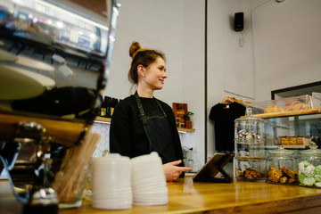 Portrait of hipster friendly female barista working with tablet while standing behind counter in modern coffee shop interior. Coffee business owner concept.