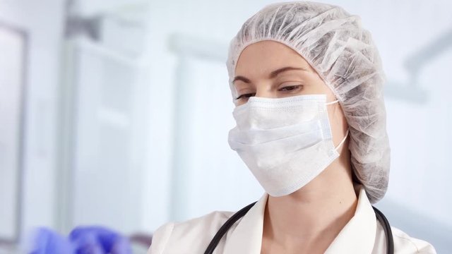 Confident professional female doctor in mask and cap in hospital room putting blue medical gloves on. Woman physician at work. Health care concept. Laboratory employee