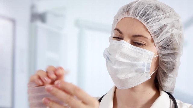 Professional female doctor in mask, cap and gloves standing in hospital room holding ampoules with new drug treatment. Woman physician at work. Laboratory employee making scientific research