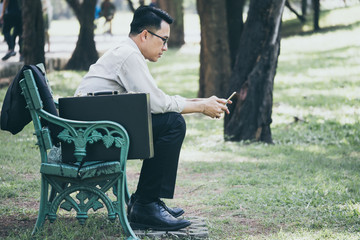 Asian businessman is using a mobile phone, sitting on the bench in the park after work hard.