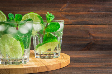 Mojito with mint and lime in a glass and a jug on the round board. Brown wood background