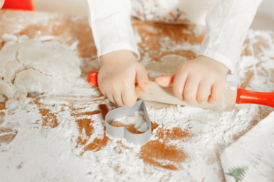 Unrecongnizable small child rolls up pastry with rolling pin, surrounded with cookie cutter in form of heart, going to bake delicious cakes with mother, involved in culinary process. Kid makes biscuit