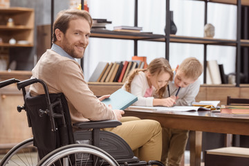 smiling father on wheelchair teaching children at home and looking at camera