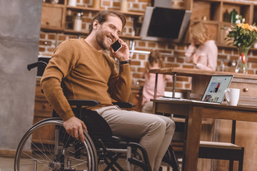 smiling disabled man in wheelchair talking on smartphone and using laptop with ebay web site at home