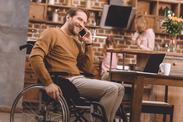 smiling disabled man in wheelchair talking on smartphone and using laptop with blank screen at home