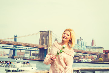 Welcome you. European Woman wearing knitted turtleneck, loose sweater, holding white rose, standing by river under sun, looking around. Brooklyn Bridge, boat with many people on background.