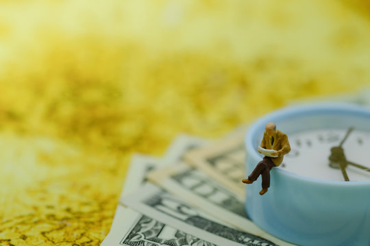 Business Money and reading concept. Businessman miniature figure sitting and reading a book on vintage round clock on US dollar bill banknotes with world map as background