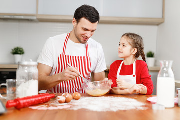Obraz na płótnie Canvas Curious female pretty child helps her father who mixes eggs, sit at kitchen, have pleasant conversation, enjoy togetherness. Little kids and her male parent bakes something delicious for party.