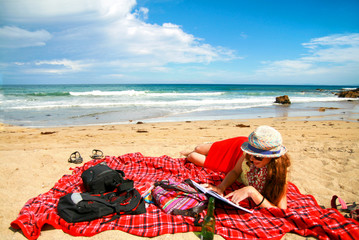 Outdoor scenery with pretty young woman in skirt laying on blanked, relaxing and reading the book at stunning, wild sandy beach