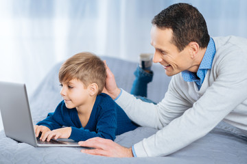 Love you. Happy middle-aged man lying on the bed next to his son and patting his head gently while the boy typing something on the laptop keyboard