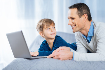 Under supervision. Happy pleasant young man lying on the bed next to his little son and watching him study online while the boy looking at him and showing the progress
