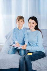 Mother-son bond. Pleasant young woman and her little son sitting on the bed together and posing for the camera while sticking memos to the notebook