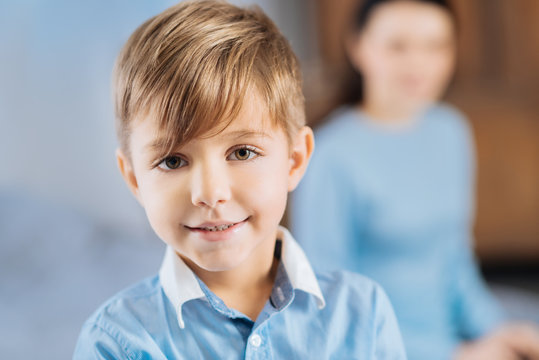 Adorable kid. The focus is on the face of a charming fair-haired little boy in a blue shirt smiling at the camera while his mother sitting behind him in the background