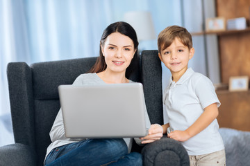 Nice pastime. Upbeat young woman and her little son watching a video on the laptop together and posing for the camera