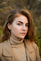 Stylish and beautiful red haired girl posing, walking in autumn forest among yellow grass. Woman with long hair, in jeans, blouse and brown coat, smiling, relaxing outdoors.