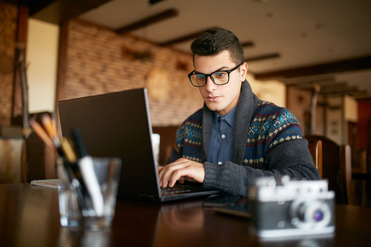 Young Attractive Hipster Man In Glasses And Winter Sweater Working On Laptop In Cafe. Freelancer Businessman Browsing Internet At Coffeshop. Photographer Retouching Photos. Vintage Camera On Table.