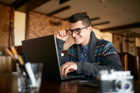 Smiling attractive hipster man in glasses and winter sweater working on laptop in cafe. Freelancer businessman browsing internet at coffeshop. Photographer retouching photos. Vintage camera on table.