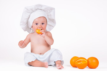 A small child eats an orange slice in a chef suit on a white background.