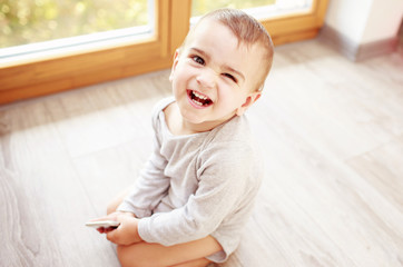Fototapeta premium Little cute boy having fun at home. Toddler in grey body seating on the floor near the window