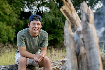 Portrait of smiling young man sitting on log at campsite during sunset