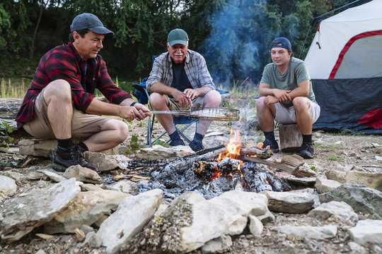 Mid Adult Man Cooking Fish On Campfire While Sitting With Friends At Campsite During Sunset