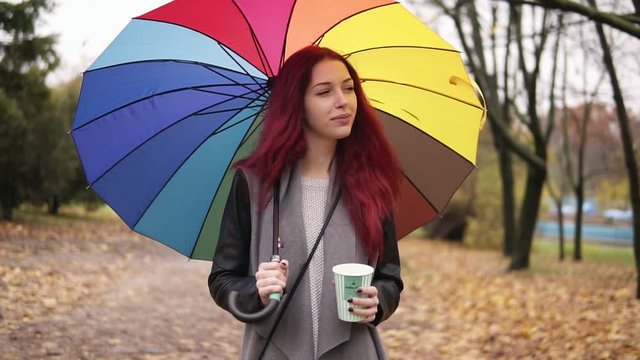 Young woman with red hair walking in autumn park and drinking coffee from a paper cup while holding colorful umbrella. Girl in warm coat enjoying cool fall weather with a cup of hot drink