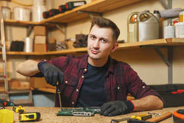 Multimeter for electrician. Caucasian young man in plaid shirt, black T-shirt digital electronic engineer repairing, soldering computer PC motherboard in workshop at wooden table with different tools.