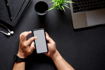 Cropped shot of man hand holding smartphone showing blank screen over  dark desk with supplies.