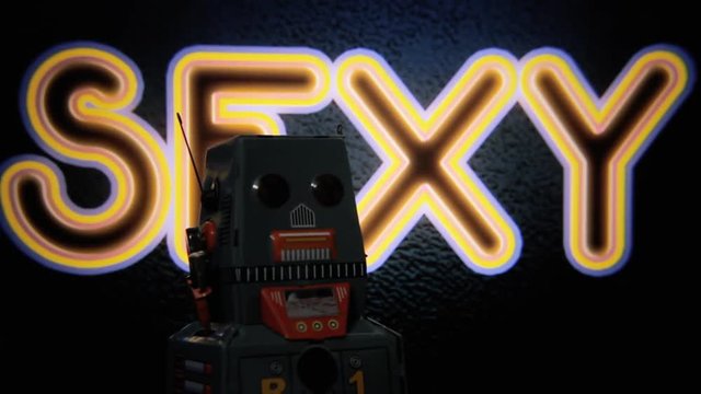 A tin toy robot moving in front of a flickering neon sign over a wall, with the text Sexy.
