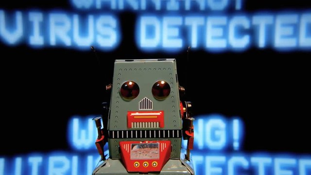 A tin toy robot moving in front of a computer screen with a looping message, Warning - Virus Detected, until it crashes down the floor.
