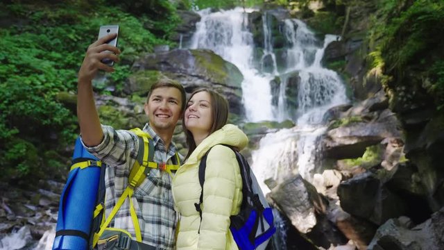A young couple of travelers photographing themselves against the background of a waterfall in the mountains. Crane shot