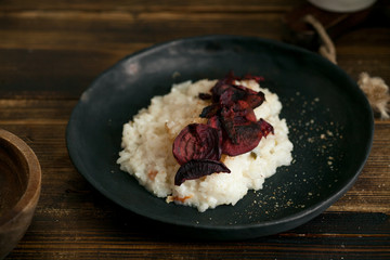 Traditional creamy tender risotto with spicy breadcrumbs and baked savoury beetroot slices in black ceramic plate on wooden table. Horizontal image, side view