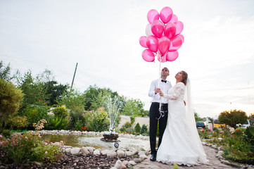 Lovely wedding couple holding a bunch of pink balloons outdoor next to the pond.