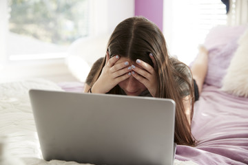 Upset teen girl lying on bed with head in hands using laptop