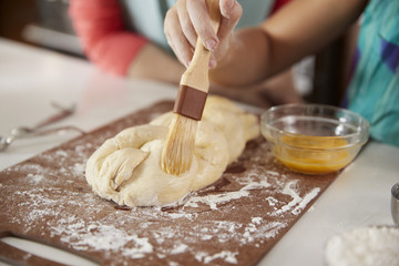 Jewish girl glazing plaited challah dough with mum, close up