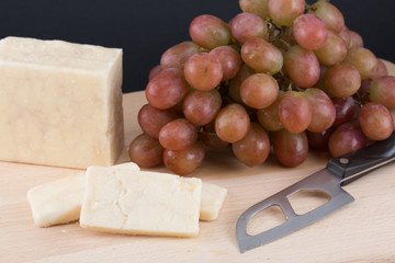 Aged cheese with a bunch of grapes and a cheese knife on a wooden cutting board on a black background