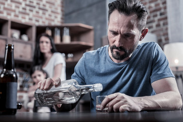 Drinking father. Serious unhappy sad man sitting at the table and pouring vodka into his glass while having alcohol addiction - obrazy, fototapety, plakaty