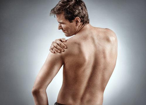 Man suffering from a shoulder injury. Photo of man holding his injured shoulder on grey background. Medical concept.