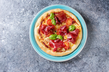 Traditional pizza with bresaola, cheese, tomatoes and basil served on turquoise plate over blue texture background. Top view with space