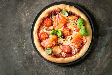 Traditionele pizza met gerookte zalm, kaas, tomaten en basilicum geserveerd op zwarte plaat over oude donkere metalen achtergrond. Bovenaanzicht met ruimte. Rustieke stijl