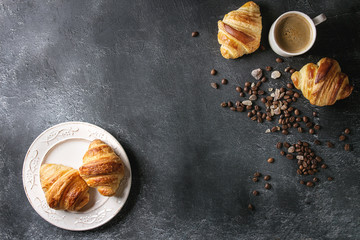 Fresh baked traditional croissants on white vintage plate and mug of espresso coffee, coffee beans, sugar over black texture background. Top view, copy space
