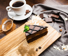 A piece of chocolate dessert on a wooden board and a gray napkin with a cup of coffee