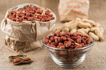 Glass bowl and paper bag full of peeled peanuts and peanuts in nutshell on sackcloth