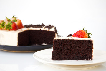 Sliced chocolate cake decorate with whipping cream, sliced chocolate and fresh strawberries. On white plate and white background.