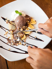 Man hands using knife and fork slice crepe with banana and chocolate icecream. French dessert. Served on white plate with whipped cream.